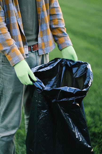 Two waste collection workers wearing dark green uniforms with reflective orange and yellow stripes are standing beside a large, industrial waste collection vehicle on an urban street. One worker is ho