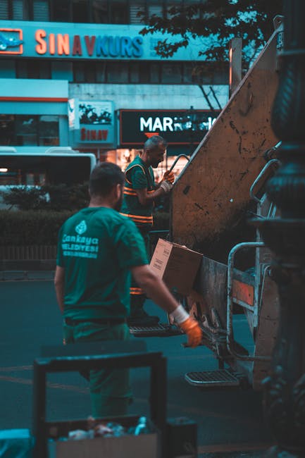 A small, blue vintage pickup truck parked on the side of a street during daytime, loaded with various types of rubbish and waste materials. The open cargo bed is filled with cardboard boxes, plastic b