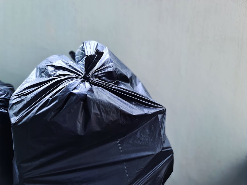 A close-up view of several tightly tied black plastic rubbish bags with a glossy finish, placed against a plain light-colored wall. The bags are stacked or leaning on each other, with the top bag prom