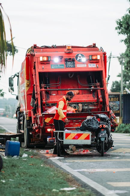 Two workers dressed in dark green uniforms with reflective orange and yellow stripes are engaged in rubbish collection on an urban street. One worker is standing on the pavement, holding a large blue 