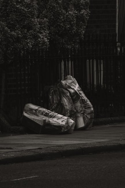 Two large black and white plastic bags filled with waste are placed on the pavement beside a curb, with one bag leaning against the other. The bags appear to contain bulky household or garden rubbish,